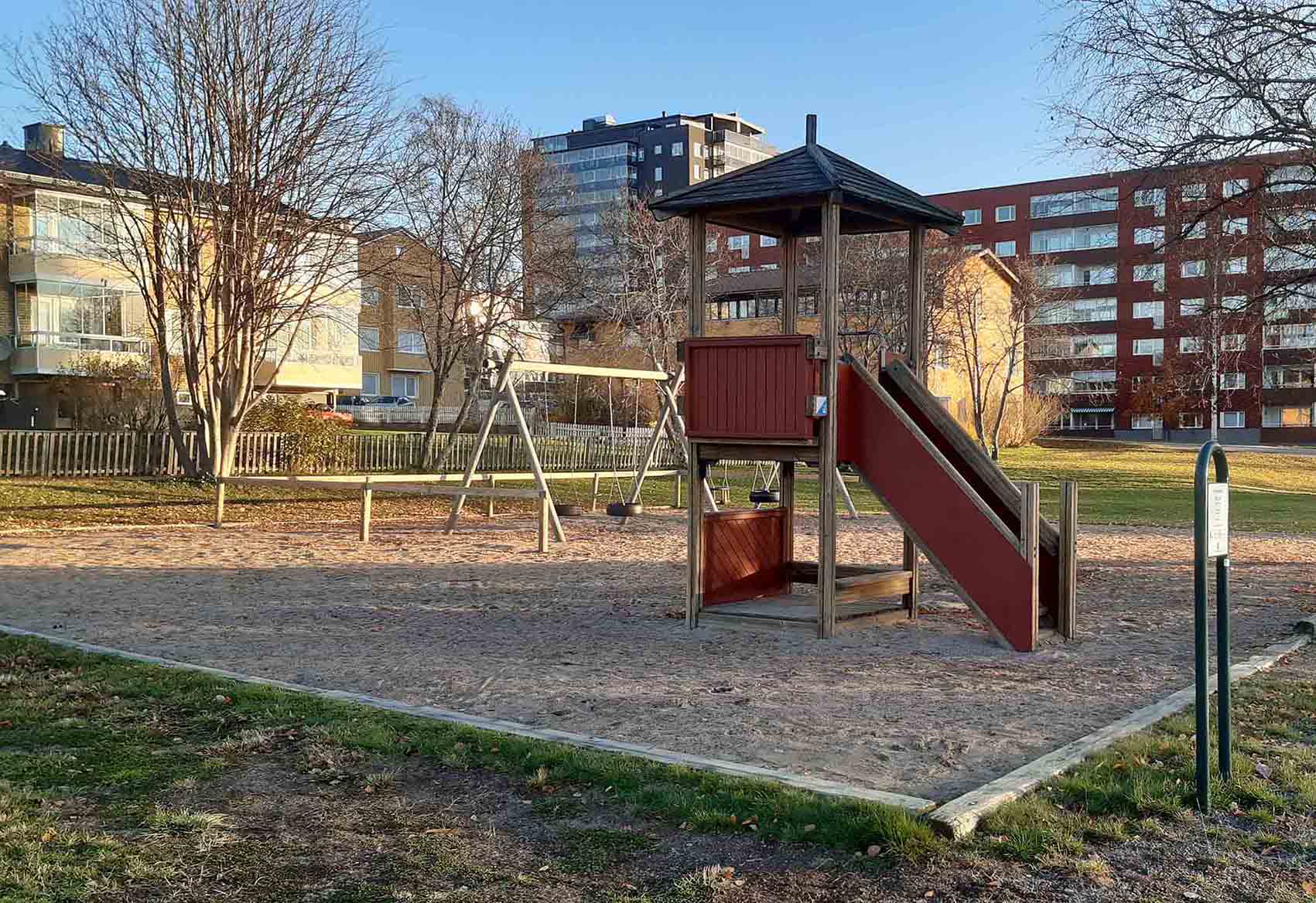 Ett lektorn och gungor på lekplatsen står på sand med avgränsat träsarg mot gräsytan runt omkring