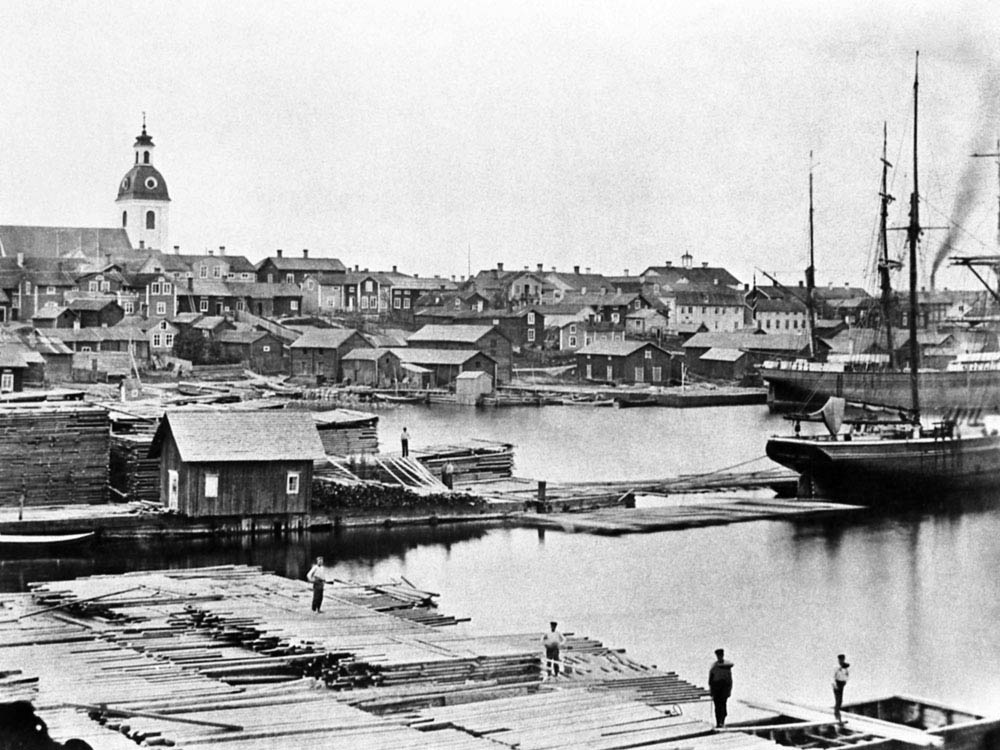 Vy över hamn och stad med många små trähus och ett segelfartyg.