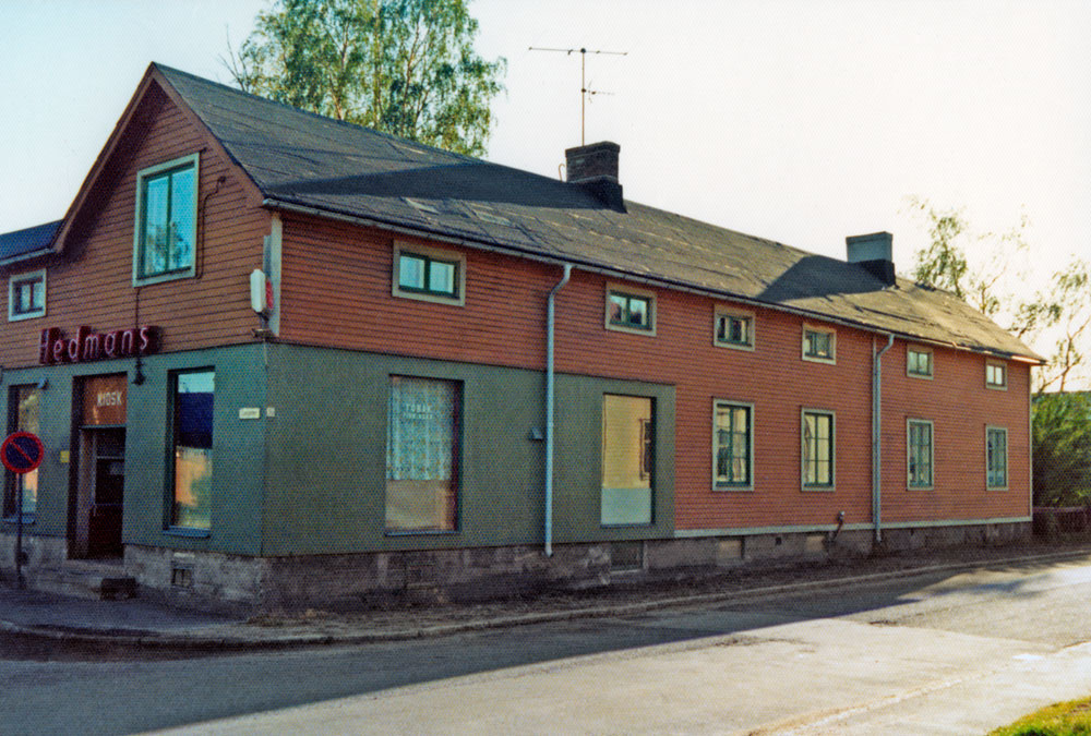 Långsträckt trähus vid gata. På gaveln ingång till butik.